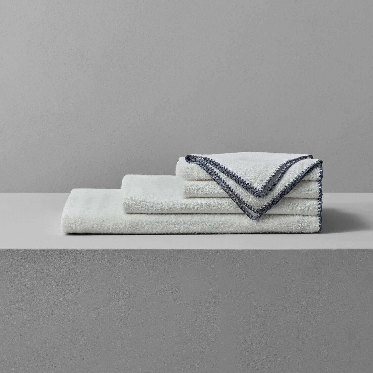 Bordado towel with grey trim, thinly plush design, eco-friendly, space-saving towels stacked on a shelf.
