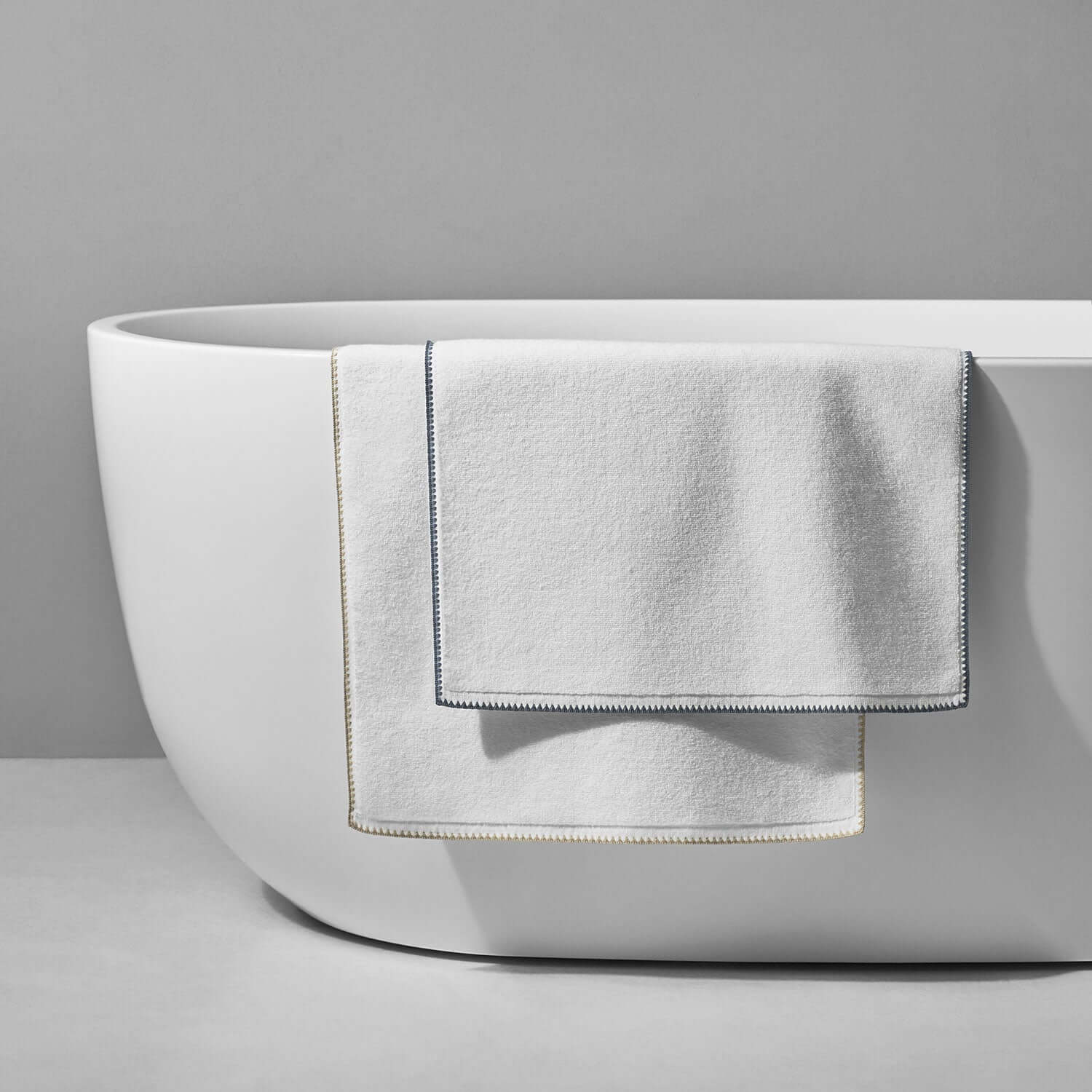 Elegant Bordado Mat gray trim towels draped over a modern white bathtub, showcasing eco-friendly design and luxury.