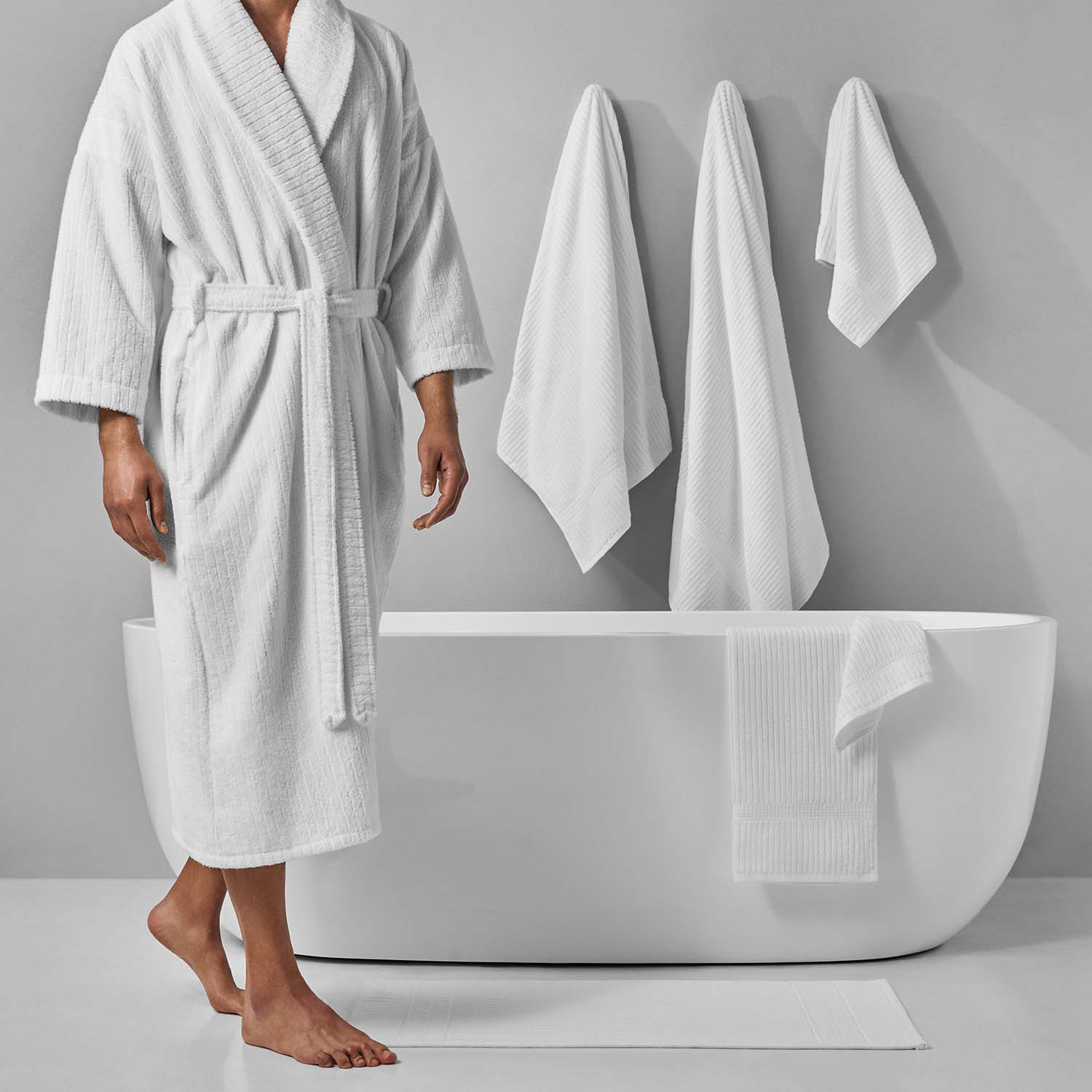 Person in a white bathrobe standing next to a modern bathtub, surrounded by fresh towels in a serene bathroom setting.