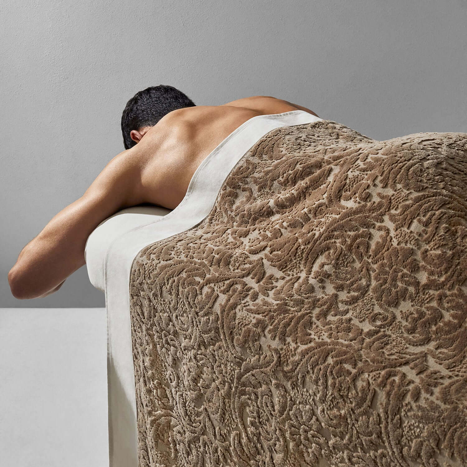 Man relaxing on a massage table covered with a luxurious Baroque Chocolate blanket, showcasing its soft velour texture.