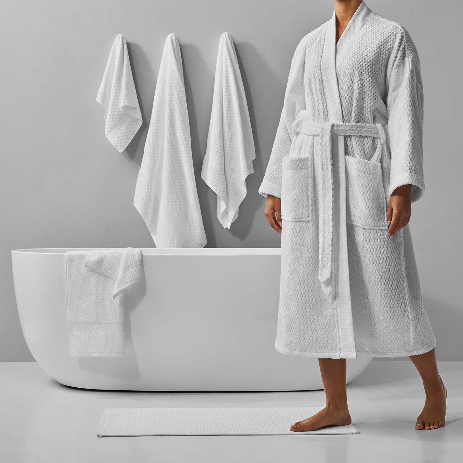 Person in a white bathrobe stepping onto a soft mat beside a minimalist bathtub with hanging towels.