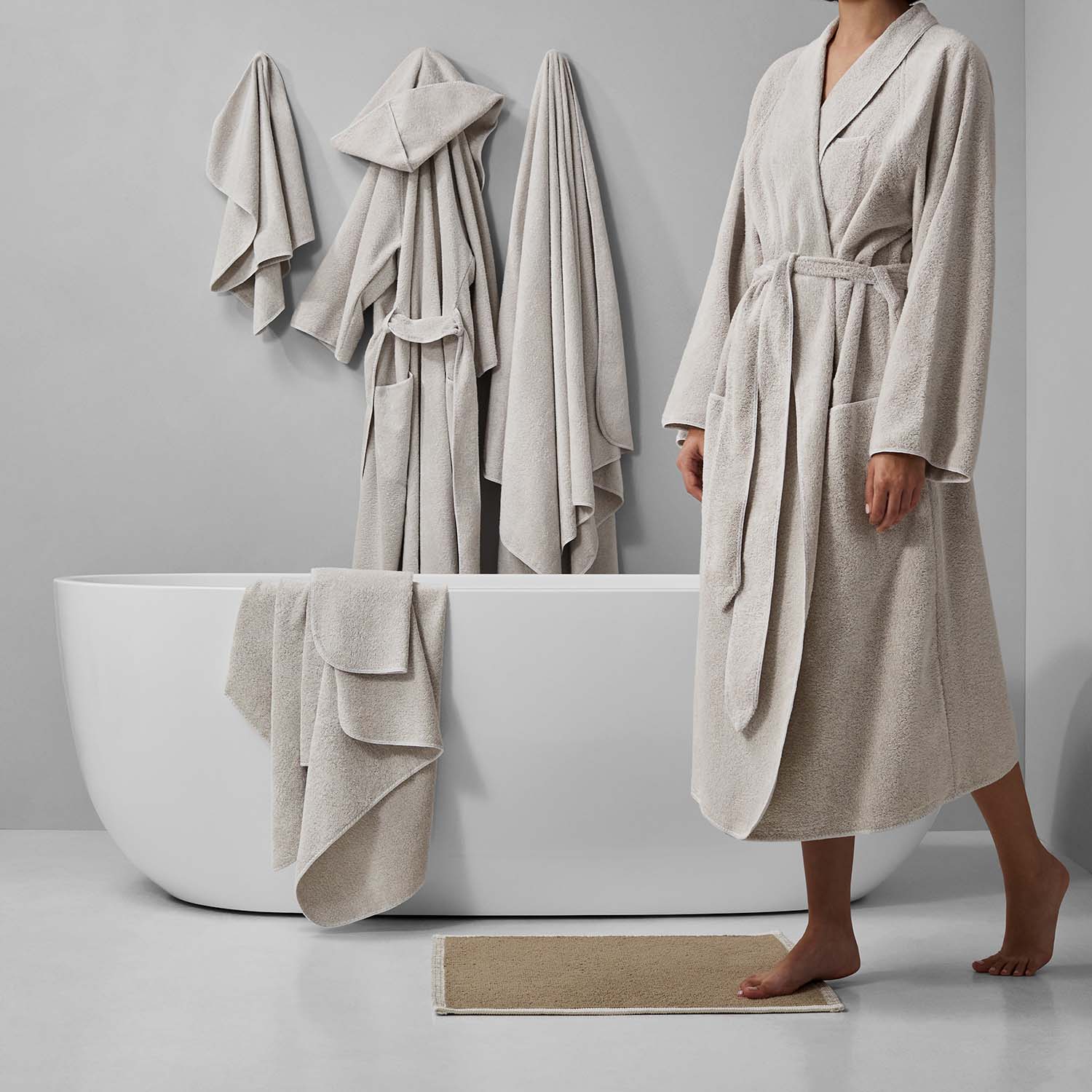 A person in a beige bathrobe stands beside a modern tub with neatly draped towels, evoking a serene bathroom atmosphere.