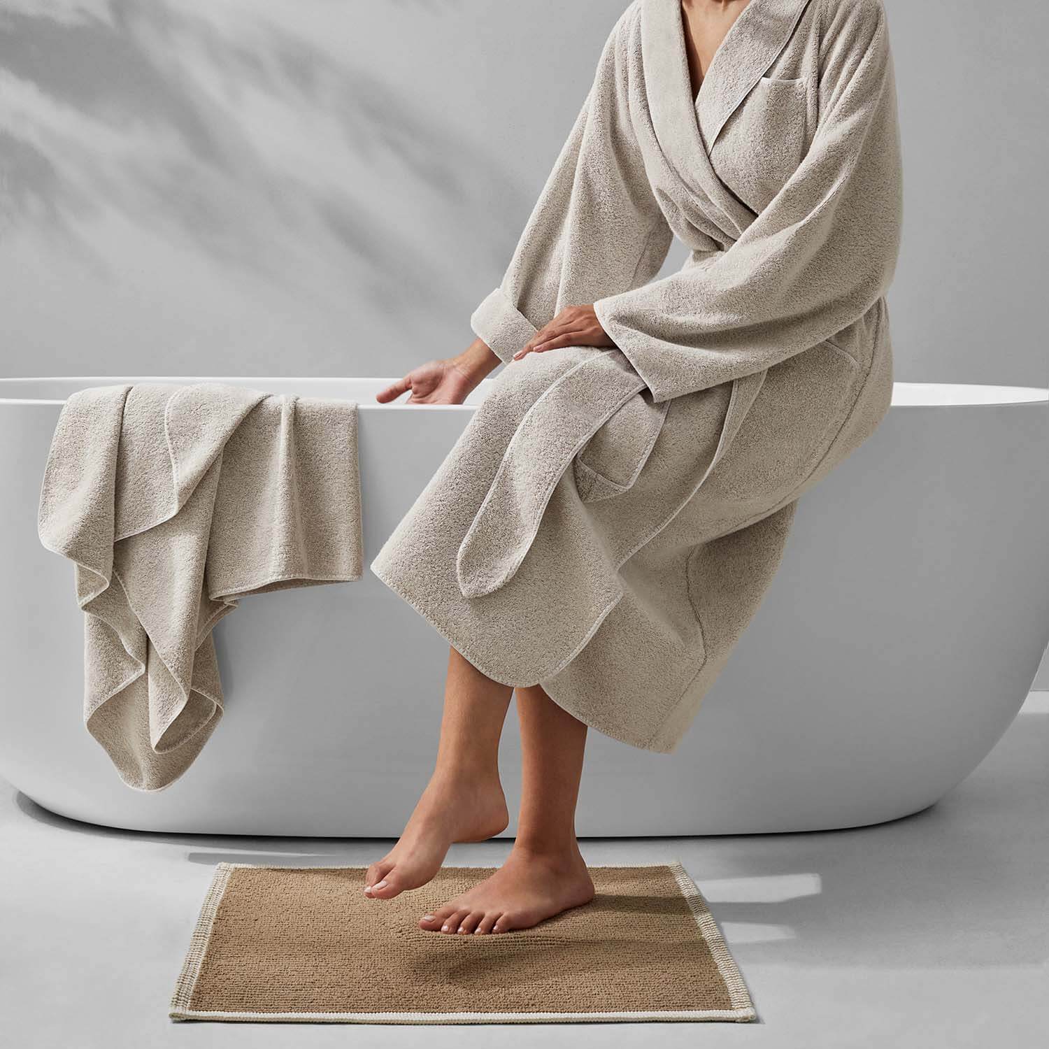 Woman in a plush beige bathrobe sitting on the edge of a modern bathtub, with towels nearby.